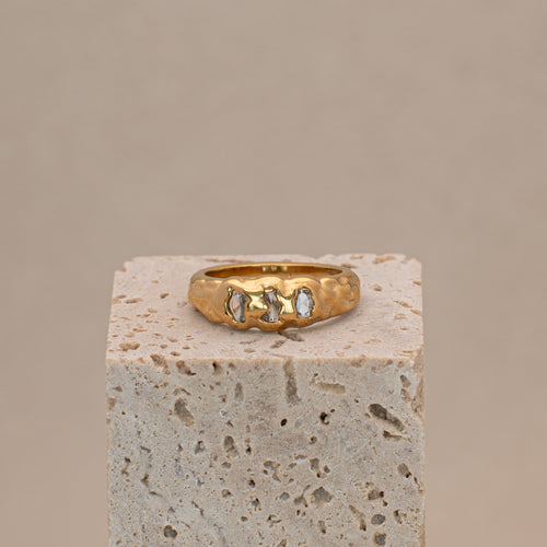 Gaia Ring in gold mit organischer Oberfläche und drei eingefassten Steinen, liegend auf einem Display.