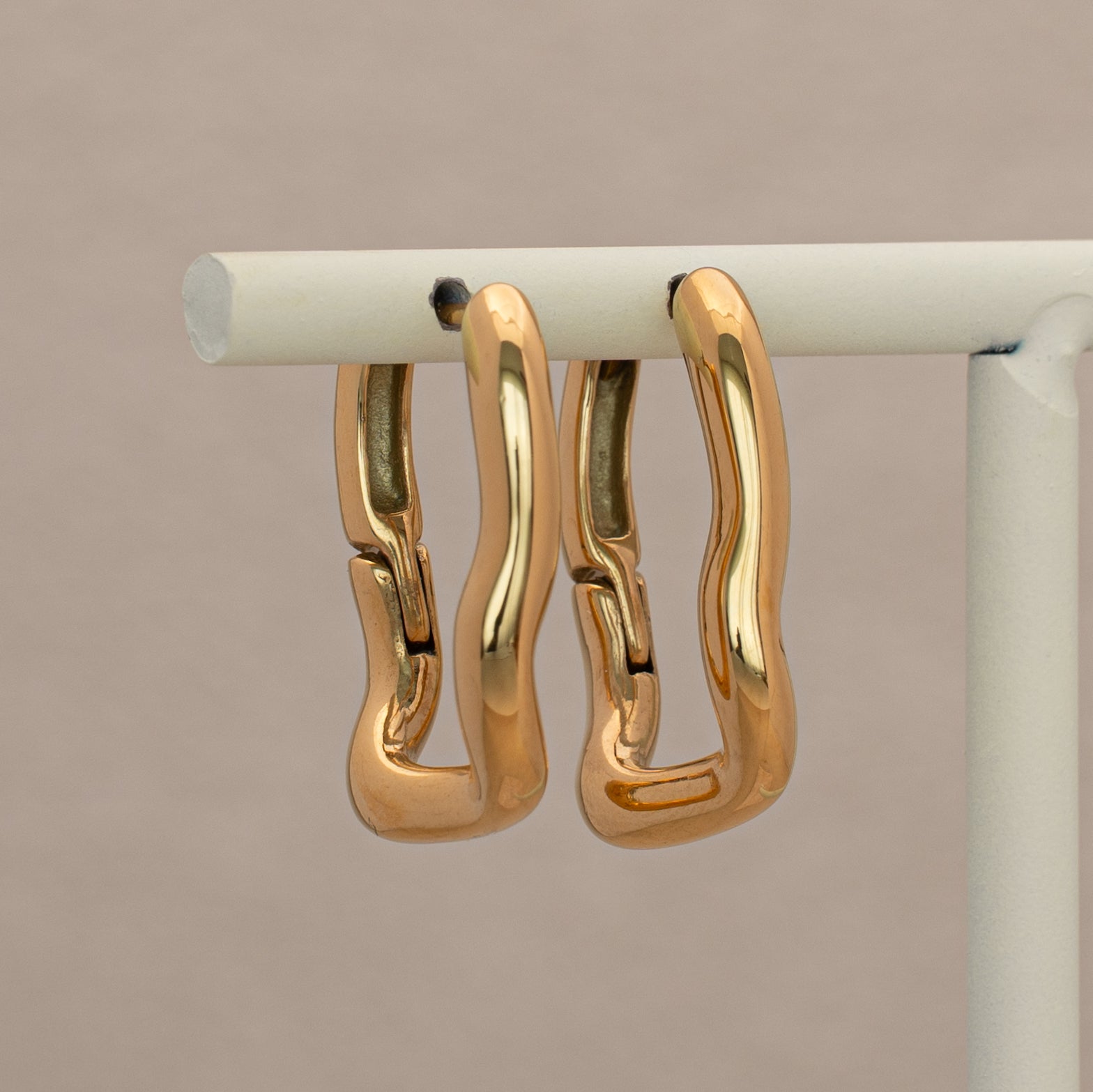 Ruwa Creolen in gold, rechteckig mit einer gewellten Form. Hängend an einem Display.